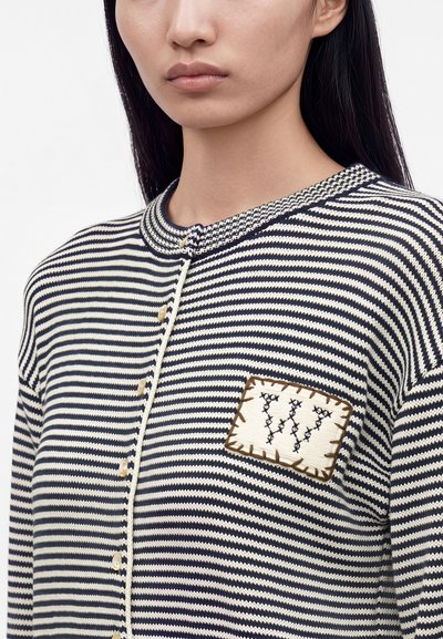 Femme portant un pull à rayures bleu marine et blanc avec des boutons, avec un écusson cousu sur la poitrine portant la lettre W.