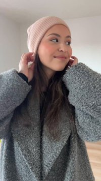 Grå fleecefrakke med brede slag, kombineret med en lyserød beanie. Personen har langt hår og justerer beanie-huen med hænderne.