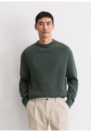 Green ribbed knit sweater with a crew neck, paired with light beige ribbed trousers featuring a button closure and side pockets.