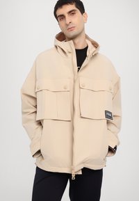 Beige, oversized jacket with a zip front, large buttoned chest pockets, and a hood. Soft fabric with a textured finish and logo patch.