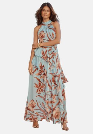 Kobieta ubrana w niebieską, bezrękawnikową sukienkę maxi z brązowym kwiatowym wzorem i falbankowymi warstwami, stoi ze skrzyżowanymi ramionami, mając na sobie sandały na wysokim obcasie.