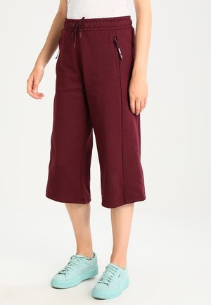 Frau trägt burgunderrote weite, verkürzte Hosen mit Kordelzug in der Taille und hellblaue Sneakers, steht vor einem einfarbigen Hintergrund.