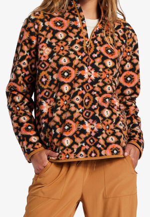 Person iført en multifarvet, luftig fleece-pullover med orange, sort, pink og hvide cirkelmønstre, kombineret med beige bukser, hænderne i lommerne.