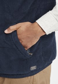 Jūras spēku krāsas kordūrveida veste ar ribotu tekstūru, sānu rāvējslēdzēju kabatu un metāla zīmola detaļu. Balta garām piedurknēm krekla piedurkne redzama pie plaukstas.