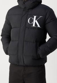 Doudoune noire avec un col montant, arborant un grand logo blanc "CK" sur la poitrine et des poches latérales. Texture lisse et matelassée.