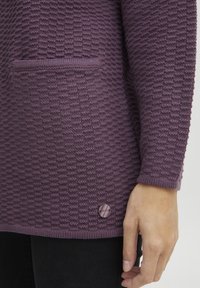 Dlouhý fialový svetr s texturou, kapsou, ribbed manžetami a dekoračním knoflíkem na lemu. Tkaný vzor je výrazný.