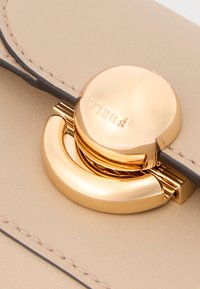 Μπεζ δερμάτινη τσάντα με στρογγυλό χρυσό κούμπωμα που φέρει το όνομα της μάρκας "PURI". Ομαλή υφή και αντίθετα σκουρόχρωμα ράμματα λεπτομέρειες.