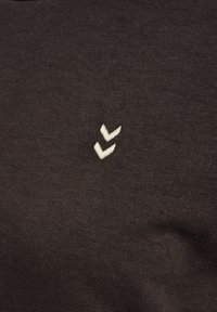 Brązowy t-shirt o miękkiej fakturze, z małym białym logo w kształcie dwóch chevronów po lewej stronie, widok z bliska.