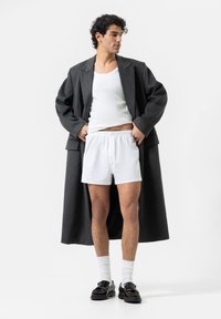 Dark gray long coat worn over a white tank top and white boxer shorts, paired with black shoes and white socks, standing against a plain background.