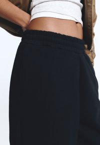 Close-up van een persoon die zwarte sweatpants met elastische tailleband draagt en een cropped wit topje met een bruine jas, hand op de heup.