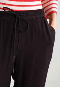Pantalons noirs en texture avec cordon de serrage et taille élastique, portés avec un haut à rayures rouge et blanc à manches longues, main dans la poche.