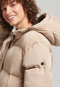 Beige pufferjakke med hætte, med et quiltet design og sort accentknap på ærmet. Glat tekstur og let materiale.