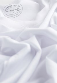 Bílá bavlněná trička s hladkou texturou a přišitou etikou, na které je v šedém písmu uvedeno "SUPERIOR QUALITY" a "100% SUPIMA COTTON".