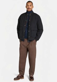 Czarna pikowana kurtka ze stójką, dwie przednie kieszenie, noszona na ciemnej koszuli, w połączeniu z brązowymi spodniami i brązowymi butami.