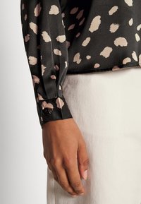 Blusa nera con macchie astratte rosa chiaro, maniche lunghe e polsini con bottoni. Tessuto strutturato, indossata sopra una gonna chiara e strutturata.