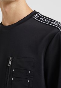 Maglietta nera a maniche lunghe con tasca con cerniera, scollatura a coste e logo bianco a contrasto sulla spalla. Tessuto liscio.