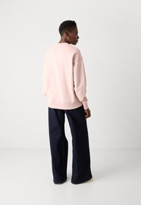 Lys rosa genser med ribbestrikkede mansjetter og hem, paret med mørke vide denimbukser. Modellen sees fra baksiden.