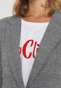 Šedé kostkované sako se strukturovaným střihem, zdobené černobílými vzory, doplněné bílým tričkem s červeným textem.