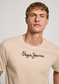 Camisa de algodón de color beige con mangas cortas. Cuenta con un escote redondo y el logo "Pepe Jeans London" en texto de color marrón oscuro y blanco.