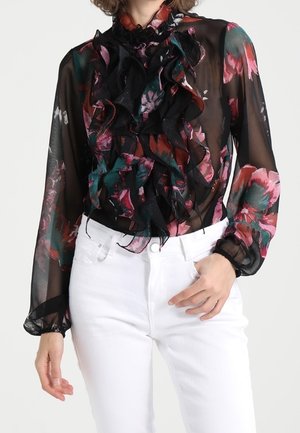 Blusa nera trasparente con motivi floreali in rosso, verde e rosa, con dettagli arricciati sul davanti e maniche a sbuffo. Indossata con jeans bianchi.