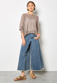 Brauner Strickpullover mit lockerem Schnitt, blaue Denim-Jeans mit weitem Bein und ausgefranstem Saum sowie braune Sandalen mit Schnallenakzenten.