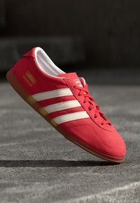 Röda mockaskor med tre vita ränder, gummiyttersula och snörningsdesign. Har logotypen "adidas" och "Gazelle" på sidan.