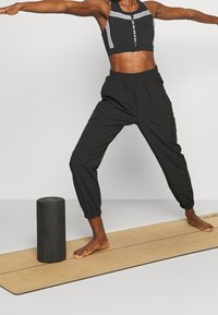 Musta lyhyt urheilurintaliivi valkoisilla raidoilla ja mustat jogger-housut. Musta sylinterimäinen vaahtorulla beigen joogamaton päällä.