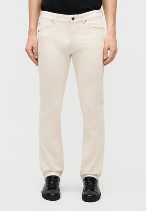 Άνδρας που φοράει μπεζ τζιν με ευθείες γραμμές και τσέπες μπροστά, σε συνδυασμό με μαύρα αθλητικά παπούτσια χαμηλού τύπου και μαύρο τοπ.