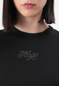 Melnas kokvilnas T-krekls ar īsām piedurknēm, kas rotāts ar spīdīgu rhinestone dizainu, kas veido vārdu "HUG". Gluda tekstūra un klasiskā apaļā apkakle.