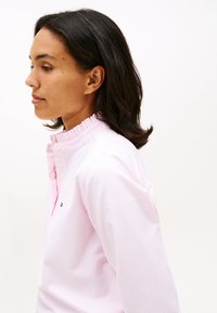 Roza in bela majica z melirano ovratnikom in nabranimi detajli; logotip vezen na levi strani prsnega koša; gladka tekstura tkanine.