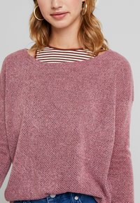 Lichtroze gebreide trui met een losse pasvorm, textuurpatroon en een brede halslijn, gedragen over een gestreept bruin-wit topje.