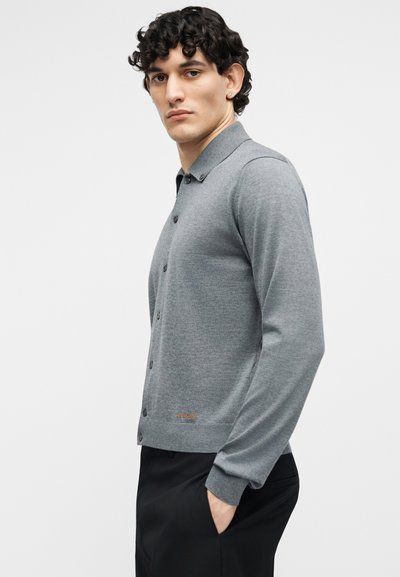 Jeune homme aux cheveux bouclés portant un cardigan gris boutonné et un pantalon noir, debout avec une main dans la poche devant un fond uni.