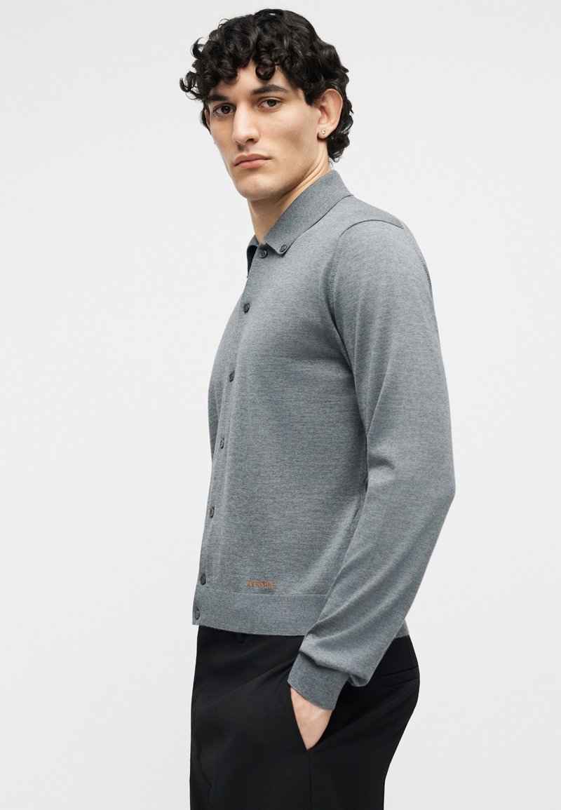 Jonge man met krullend haar, gekleed in een grijze gebreide vest met knopen en een zwarte broek, staand met één hand in de zak tegen een effen achtergrond.