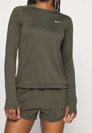 Persoon draagt een olijfgroen Nike-shirt met lange mouwen en bijpassende korte broek, met zichtbare Nike-logo's op de borst en shorts.