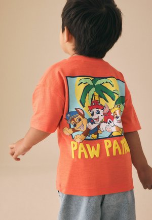 Vaikas vilki oranžinį marškinėlį su spalvingais Paw Patrol personažais ir atspaudą su palmės medžiu ant nugaros, prie kurio priderintos šviesios džinsinės šortai.