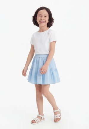 Ragazza sorridente in piedi con capelli corti e scuri, indossa una maglietta bianca, una gonna azzurro chiaro e sandali bianchi su uno sfondo bianco uniforme.