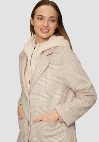 Manteau à carreaux rose clair avec un design structuré, présentant de grands revers, des poches avant et une doublure à capuche rose doux. Texture confortable dans l'ensemble.