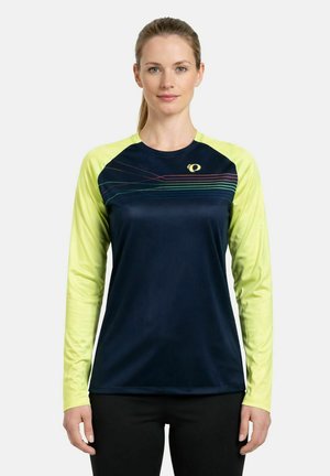 Femme portant un chemisier bleu marine à manches longues avec des manches jaunes et de fines rayures colorées sur la poitrine, debout les bras détendus.