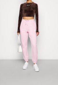 Rajoitetun pitkän hihansuin varustettu ruskea samettinen lyhyt toppi logolla, yhdistettynä vaaleanpunaisiin elastisiin nilkka-sweatpants-housuihin, joissa on sivulogopatch. Valkoiset lenkkarit ja laukku.