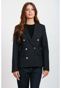 Blaser doppiopetto navy in tessuto liscio con bottoni dorati, spalle strutturate e due tasche frontali. Abbinato a jeans scuri.