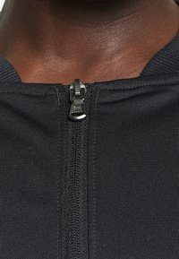Black zip-up garment with a textured fabric, featuring a metallic zipper at the neck. Close-up view of the zipper and neckline details.