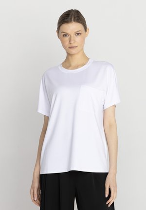 Kobieta stojąca, ubrana w prosty biały T-shirt z krótkim rękawem i kieszonką na piersi oraz luźne czarne spodnie na jednolitym jasnym tle.