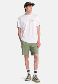 Λευκό βαμβακερό μπλουζάκι με πράσινο λογότυπο, χακί σορτς, μαύρο καπέλο, λευκές κάλτσες με μαύρες ρίγες και μπεζ αθλητικά παπούτσια με υφή λεπτομέρειας.