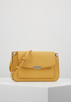 Žlutá kožená kabelka přes rameno s nastavitelným popruhem a stříbrným kovovým zapínáním, vystavená na bílém podstavci na jednobarevném pozadí.