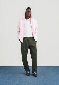 Roza majica s kapuljačom na patentni zatvarač preko bijele majice, u kombinaciji sa zelenim suženim hlačama i crnim tenisicama. Minimalistički dizajn s ležernim krojem.