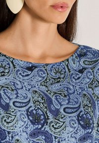 Frau mit großen weißen Tropf-Ohrringen und einem blauen Oberteil im Paisleymuster mit runder Halslinie, Lippen mit glänzendem rosa Lippenstift.