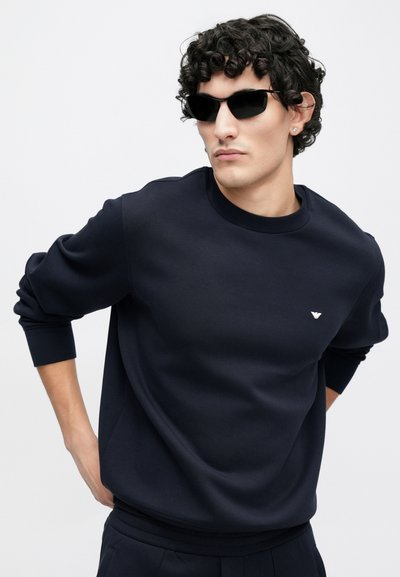 Tmavomodrá mikina s okrúhlym výstrihom a rebrovanými manžetami; na hrudníku malý biely logotyp; hladká textúra látky.