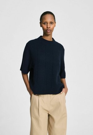 Femeie purtând un pulover scurt cu mâneci striate, de culoare bleumarin, ușor băgat în pantaloni bej plisati, cu mâinile în buzunare, stând în fața unui fundal simplu.