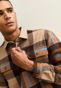 Brown and beige plaid flannel shirt with blue accents, corduroy collar, and buttoned front pockets. Textured fabric with a casual design.