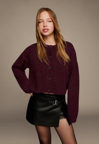 Burgunderfarbene, kurz geschnittene Strickjacke mit großen Knöpfen, kombiniert mit einem schwarzen Leder-Minisrock mit einem Gürtel-Detail. Glatte Textur.